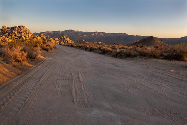 A dirt road leads towards rocks, a desert valley, and mountains.