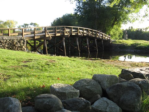 Arched wooden bridge with field stone abutments spans a slow river bounded by green embankments