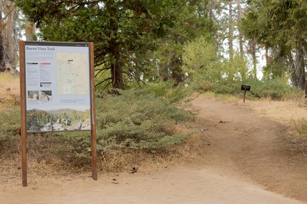 A metal framed sign stands to the side of a dirt trail. Behind the sign are bushes and trees.