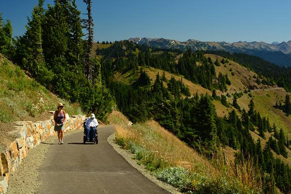 A hiker and wheelchair user on a paved trail with mountains in the background.