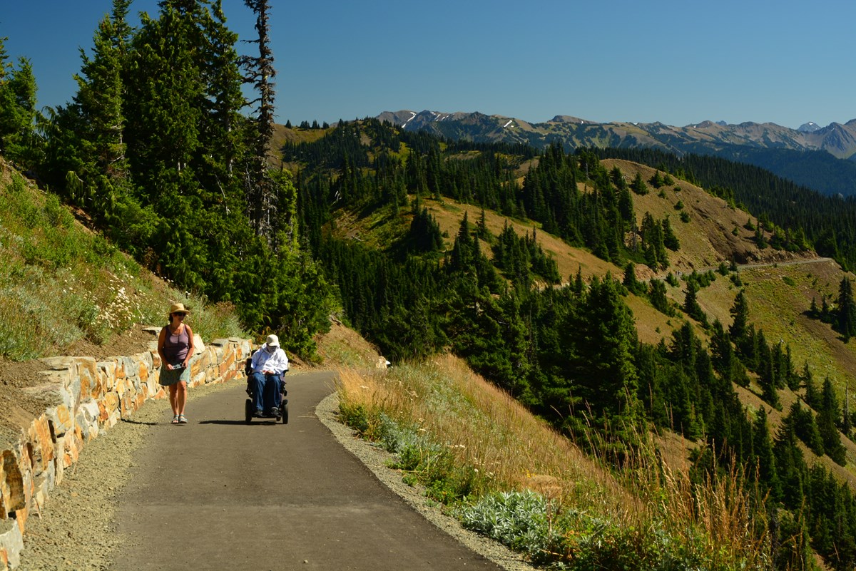 A hiker and wheelchair user on a paved trail with mountains in the background.