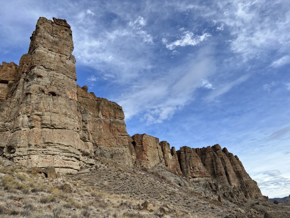 Tan cliff formations with a grassy slope below dotted with fallen boulders.