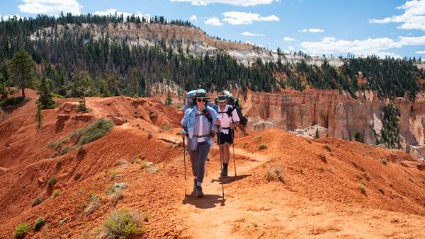 Two backpackers hike along red rock path with distant forest and cliffs