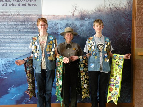 A ranger standing between two junior rangers with vests and banners covered in junior ranger badges.