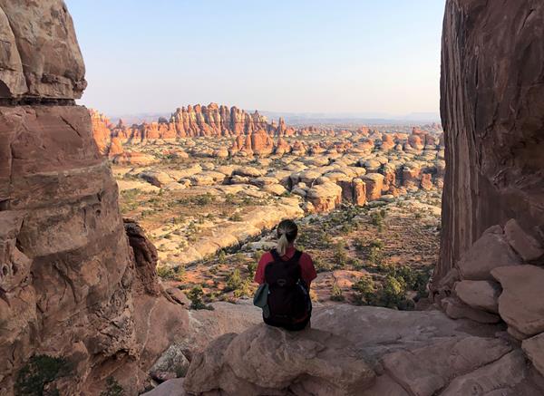 A hiker sits atop a sandstone rock looking out across a vast view of sandstone spires.