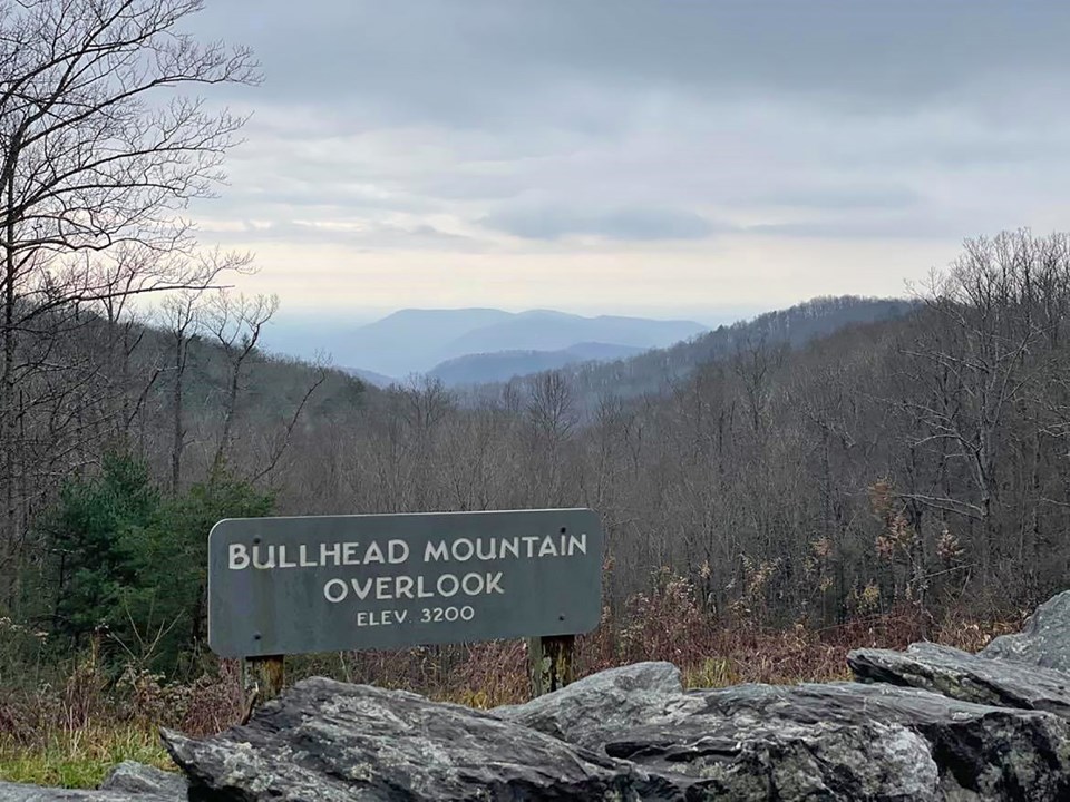 A rock wall and sign stand in front of a bare, winter forest, with mountains in the distance