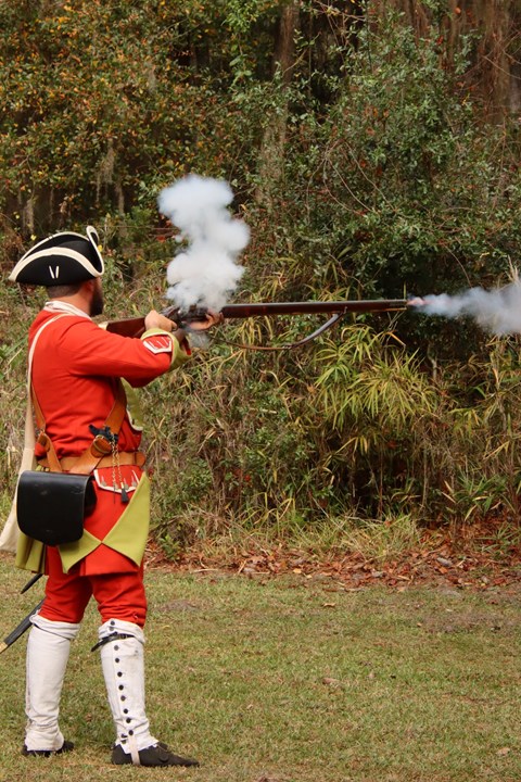 British soldier wearing colonial uniform firing a musket with smoke coming out of the muzzle.