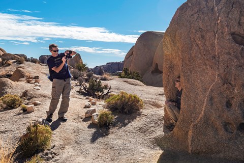 A man on a trail holds a camera and smiles towards a person kneeling in front of a rock