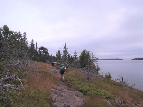 Four people walk on rocky trail towards a forest near a shoreline.