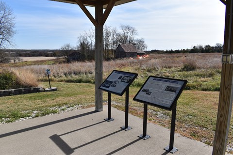 Two information signs stand at the edge of a concrete floor, which overlooks fields and old cabins.