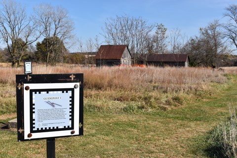An information sign with QR code stands in front of historic wood cabins in the distance.