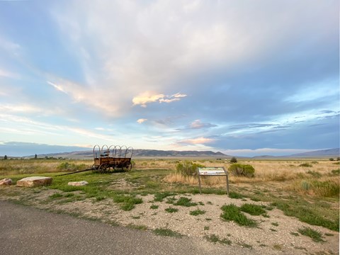 A wayside exhibit sign next to a prairie schooner wagon replica, beautiful sunset clouds.