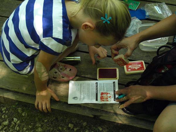 A girl bends over quest brochure as an adult’s hands stamp the back. They are on a wooden platform.