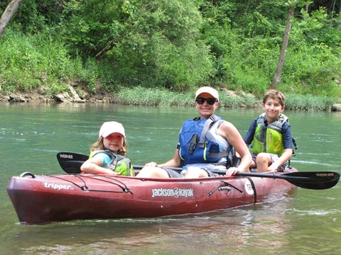 Red kayak on river with woman in middle, girl in front, boy in back, all wearing PFD