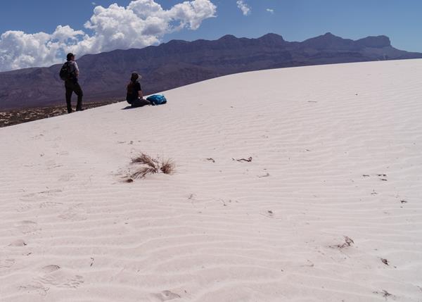 Two people stand on white sand dunes in front of a desert mountain range