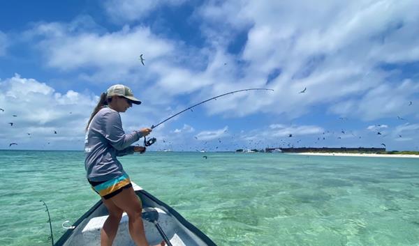 A female angler fights a fish in a small boat atop of crystal clear blue waters.