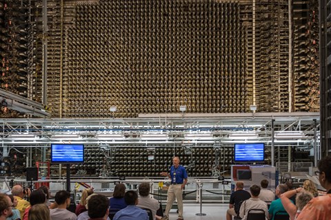 A group of people sit in chairs as a man gives a talk in front of a large industrial apparatus.