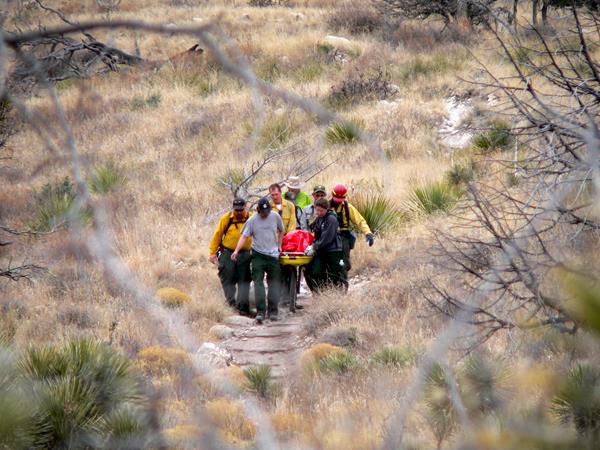 A group of uniformed staff carry a wheeled litter down a rocky trail in a desert mountain terrain