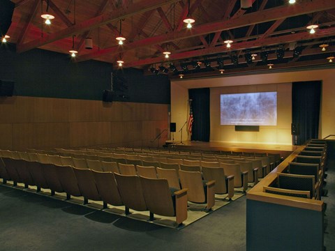 An auditorium with wood wall, exposed truss ceiling, and stage with screen.