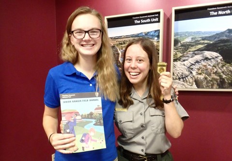 Two women smiling at the camera, one in a ranger uniform, holding a junior ranger book and badge.