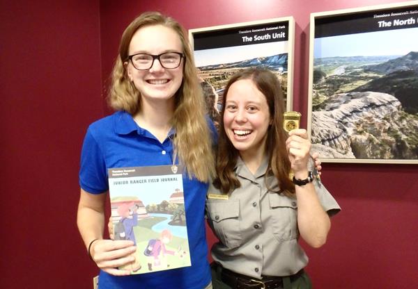 Two women smiling at the camera, one in a ranger uniform, holding a junior ranger book and badge.