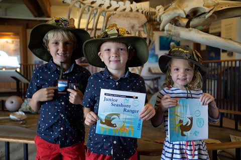 Three kids wearing ranger-like hats, smiling and holding up booklets in front of a whale skeleton