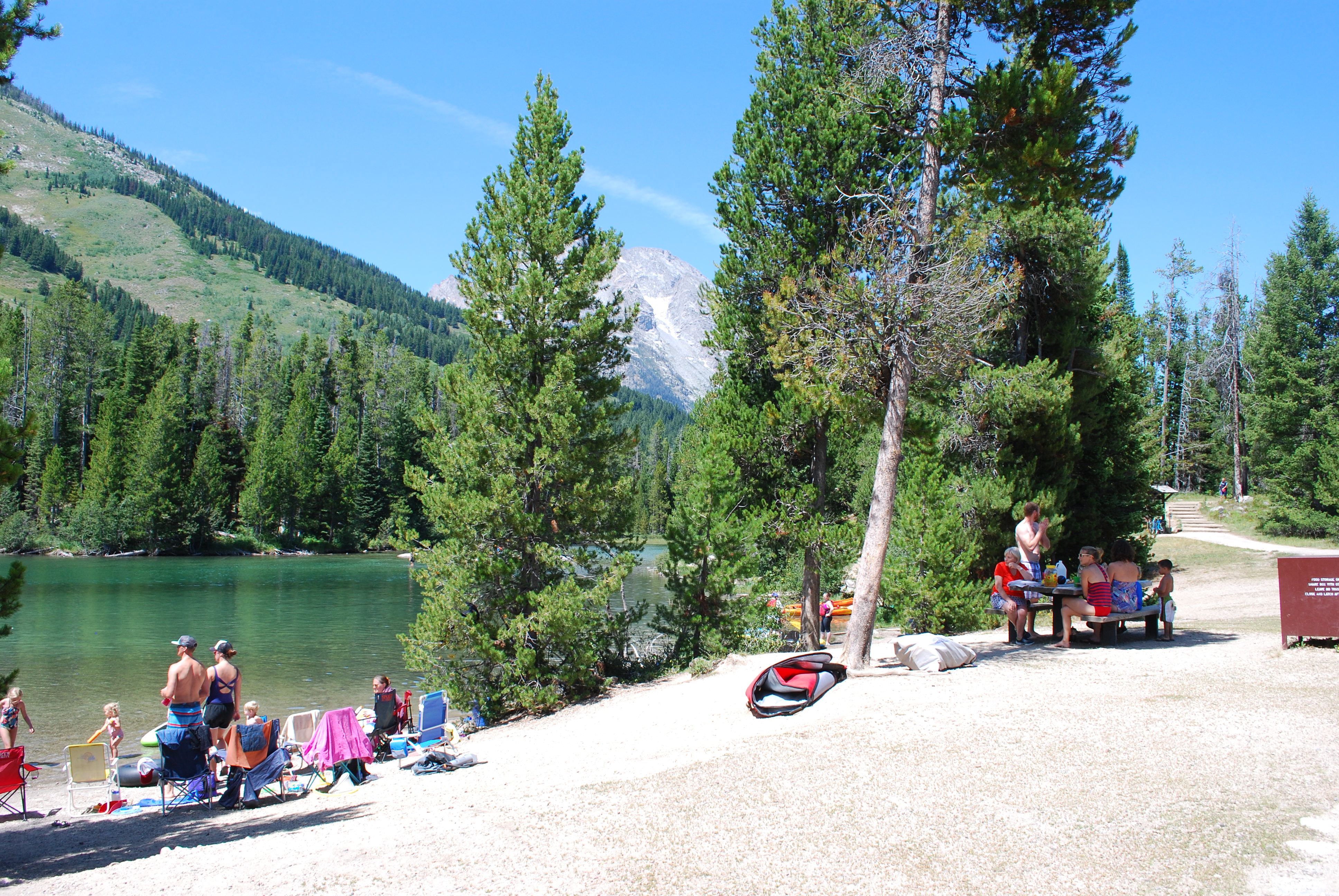 A couple different family groups sit along a lake and at a nearby picnic table.