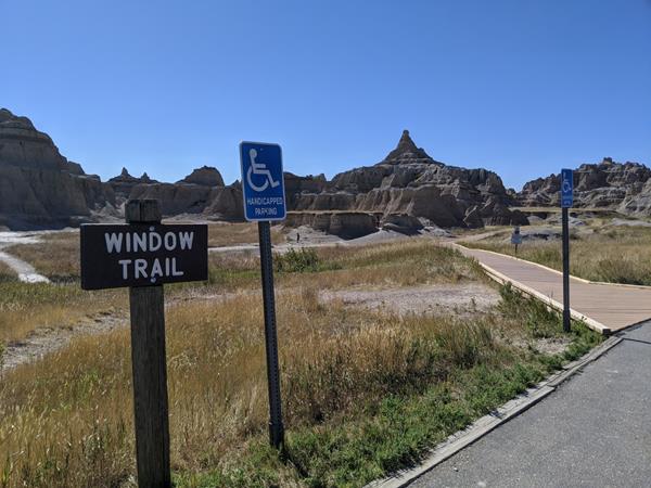 A sign marked Window Trail next to blue ADA sign with badlands buttes and boardwalk under blue sky.