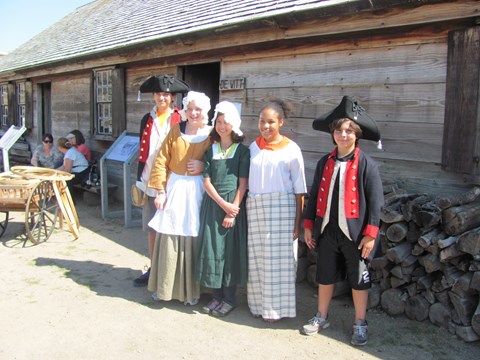 Five children stand in a row smiling. They all have different 18th Century accessories on.