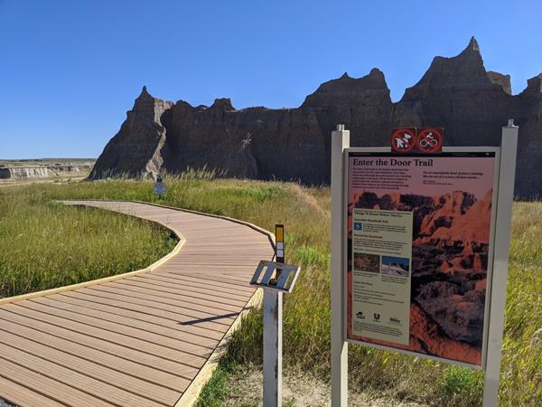 A sign marked Door Trail next to blue ADA sign with badlands buttes and boardwalk under blue sky.