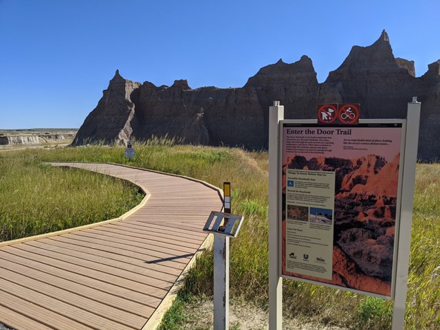 A Door Trail sign and an accessible-trail sign beside a boardwalk leading toward badlands buttes under blue sky