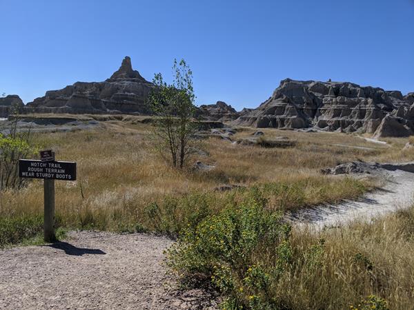 A sign marked Notch Trail alongside a trail proceeding amid badlands buttes under blue sky.