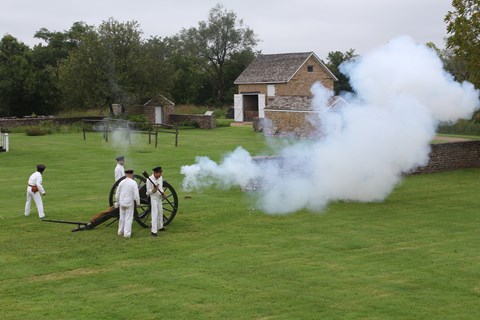 four men in white uniforms firing a cannon. Smoke is coming out of the cannon.