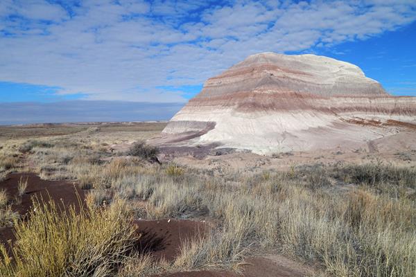 Old road winding through grassland in front of banded badlands under a blue sky with some clouds