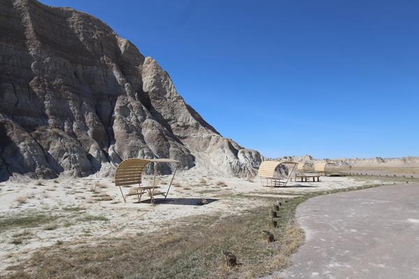 Two shaded picnic tables beside paved road, badlands butte, and under a blue sky.