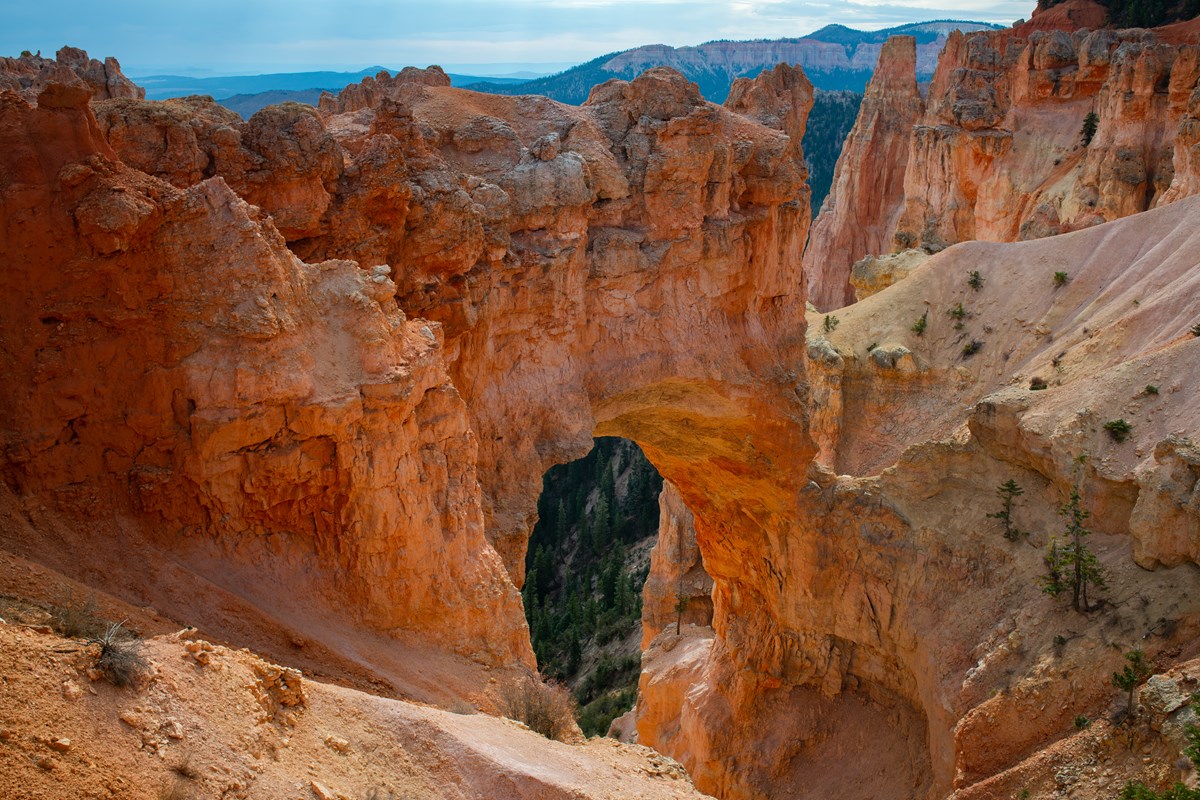 A glowing orange rock arch stands among limestone slopes above a forested landscape