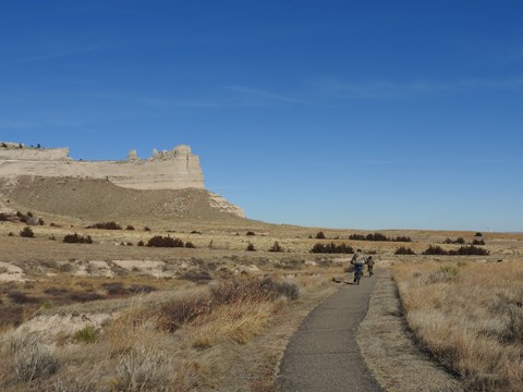 A pair of bicyclists are seen riding towards a distinctive sandstone rock formation.