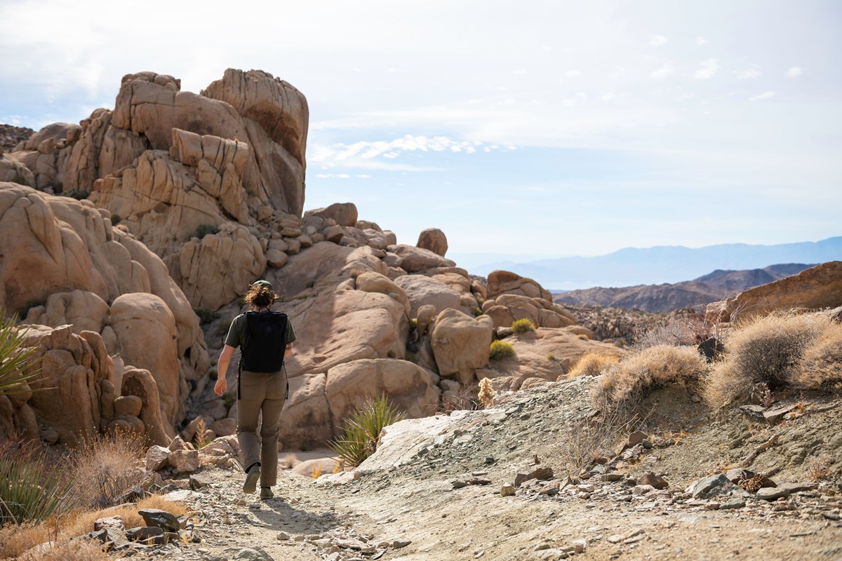 A hiker walks down a scree-covered trail next to tall boulders.