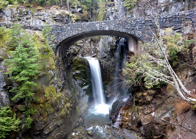 A gray stone bridge frames water cascading down a rocky ledge into a river below.