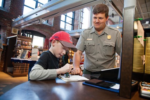 A boy stamps an activity book while a park ranger watches.