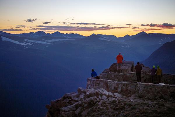 people stand on a platform watching the sun set behind mountains