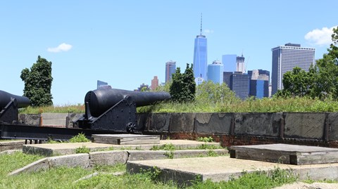 large, black, iron cannon pointing over forts wall with NYC skyline in backround