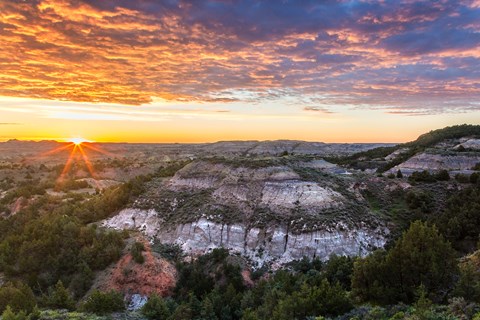 The sun is just visible over a badlands scene. Juniper grow on the buttes, and there are low clouds.
