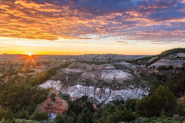 The sun is just visible over a badlands scene. Juniper grow on the buttes, and there are low clouds.