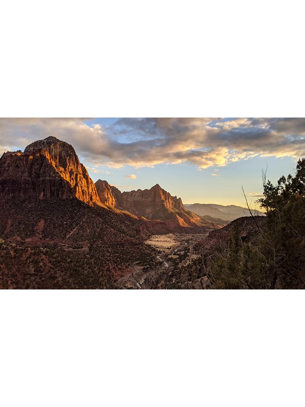 A cloudy blue sky over red sandstone mountains, with green foliage dotting their foothills.