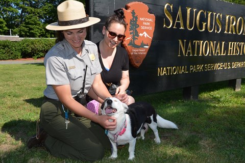 Two people sit on the grass petting a black and white dog in front of the Saugus Iron Works sign