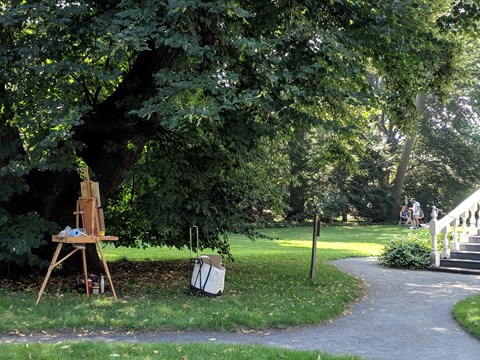 artist's easel set up under large tree, with group of people around second easel on lawn behind