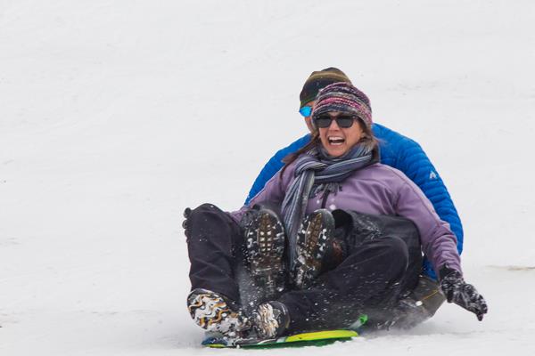 Two visitors are sitting on a sled and sledding down a snowy hill