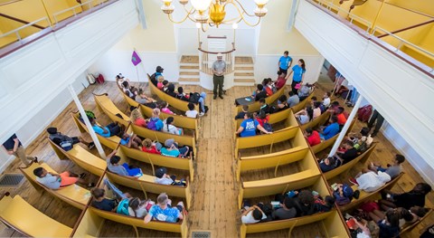 View from second story onto first floor of meeting house that has pews filled with kids.