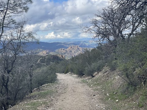 Rocky trail heading down chaparral covered mountain with rock formations seen from a distance.